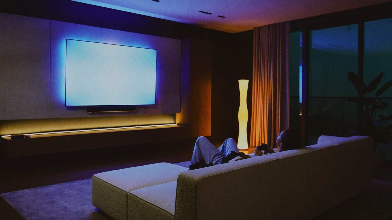 A modern open-plan lounge at night with a backlit media wall glowing softly behind the screen